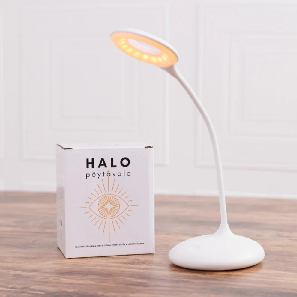 North Ray HALO Pöytävalo; moderni valkoinen pöytävalaisin pyöreällä LED-päällä hehkuu laatikon vieressä, jossa lukee "HALO pöytävalo" ja on silmäkuvitus; asetelma puisella alustalla ja minimalistisella valkoisella seinätaustalla; valaisin on sileä, mattapintainen ja kokonaan valkoinen; laatikko on suorakulmainen, mustaa groteskitekstiä ja kuvitettu silmä; ei ihmisiä tai käsiä näkyvissä, kuva hyvin valaistu.