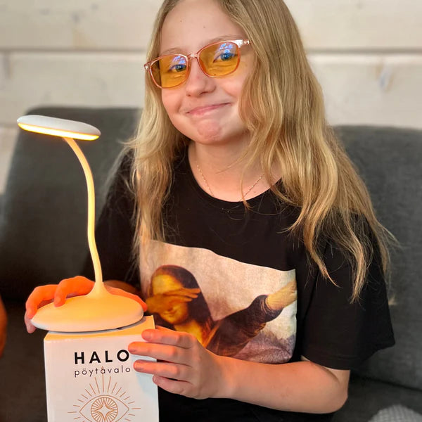 North Ray HALO Pöytävalo; hymyilevä pitkähiuksinen tyttö oranssit silmälasit päässään pitelee pyöreää Halo-pöytävalaisinta, joka säteilee kellertävänvalkoista valoa; valaisin on kapea, metallinen ja mattapintainen; tyttö istuu tummalla kangassohvalla ja hänellä on musta t-paita, jossa Mona Lisa -kuva keskellä; pakkausta tai tekstiä ei näy kuvassa.