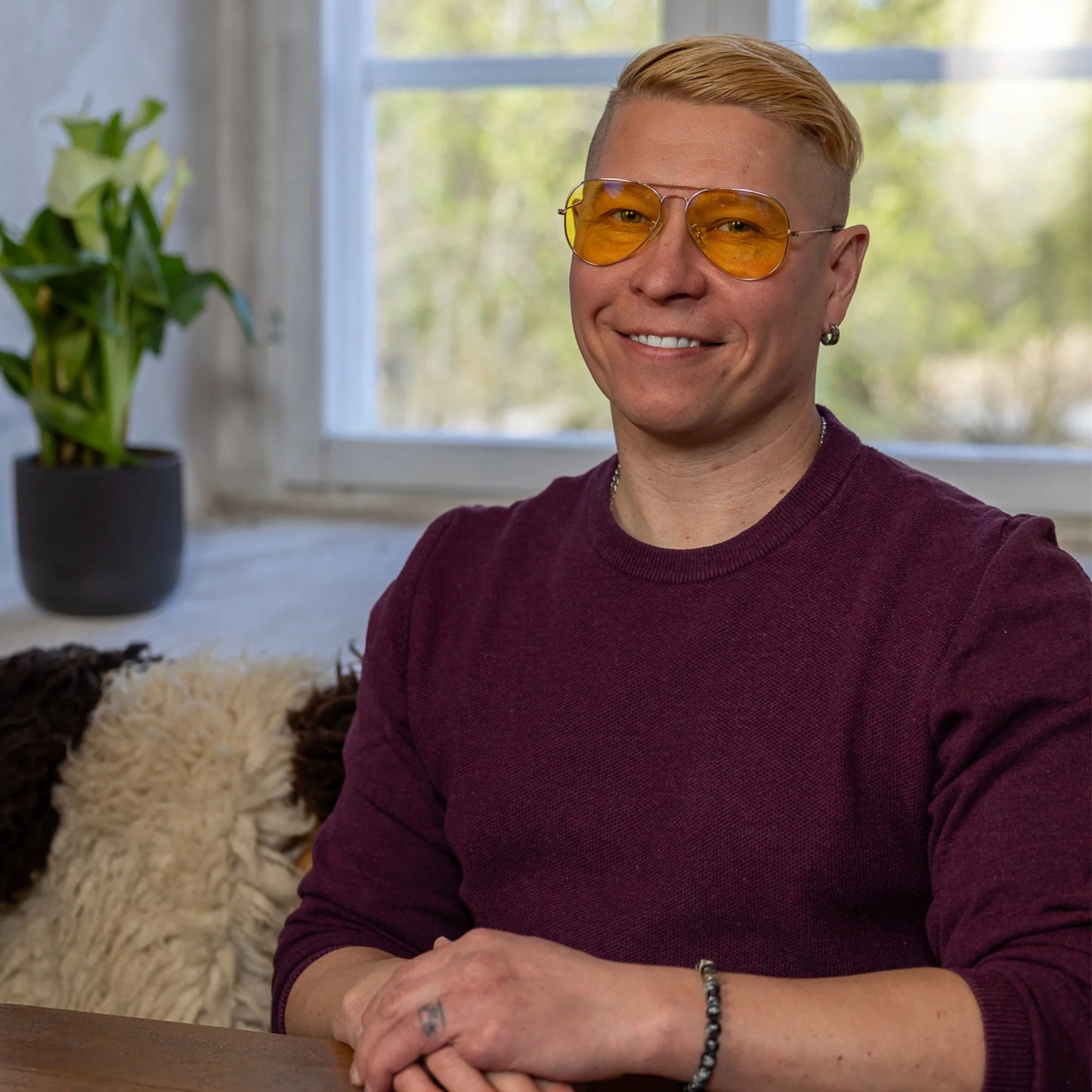 North Ray ILMARI Sinivalolasit; hymyilevä lyhythiuksinen vaaleahiuksinen henkilö keltaisissa laseissa istuu sisätiloissa ikkunan äärellä pukeutuneena viininpunaiseen neuleeseen; taustalla ruukkukasvi ja pörröinen peitto luovat kodikkaan sisätilan tunnelman; ei pakkausta, näkyviä etikettejä tai tuotebrändäystä; ei erikoisia pintakäsittelyjä tai muotoiltuja pakkauksia näkyvissä kuvassa.