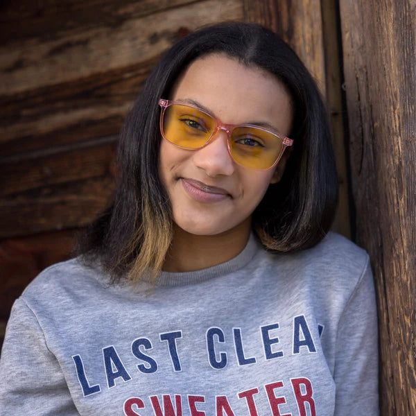 North Ray KAJO Sinivalo Päivälasit; suoratukkainen tummahiuksinen henkilö keltalaseissa hymyilee nojaten puiseen seinään; henkilöllä keskiharmaa collegepaita rinnassa versaalitekstillä "LAST CLEAN SWEATER" valkoisin lohkokirjaimin; seinä puuta pystysuorin lankuin näkyvin syinen; kuva vyötäröltä ylöspäin, ei pakkausta näkyvissä.