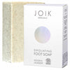 A rectangular exfoliating foot soap bar stands next to a white box labeled JOIK Organic Exfoliating Foot Soap. The soap bar is a neutral shade with textured particles visible on its matte surface. The box is white, rectangular, and has black sans-serif typography in upper and lower case. The design is minimalist, showing product name, organic certification mark, and brief details. Both items are placed on a plain background. No people or hands are present in the image. Shot is front-facing at product level.