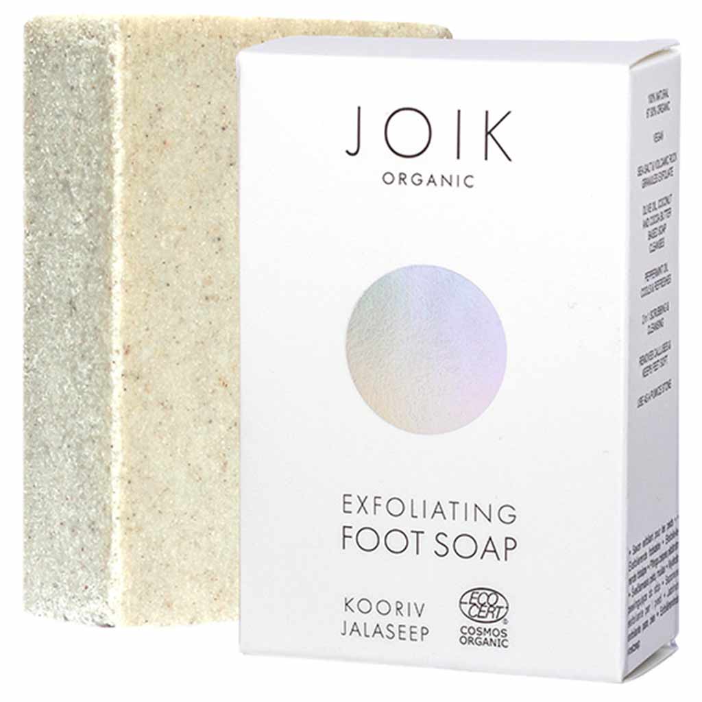 A rectangular exfoliating foot soap bar stands next to a white box labeled JOIK Organic Exfoliating Foot Soap. The soap bar is a neutral shade with textured particles visible on its matte surface. The box is white, rectangular, and has black sans-serif typography in upper and lower case. The design is minimalist, showing product name, organic certification mark, and brief details. Both items are placed on a plain background. No people or hands are present in the image. Shot is front-facing at product level.
