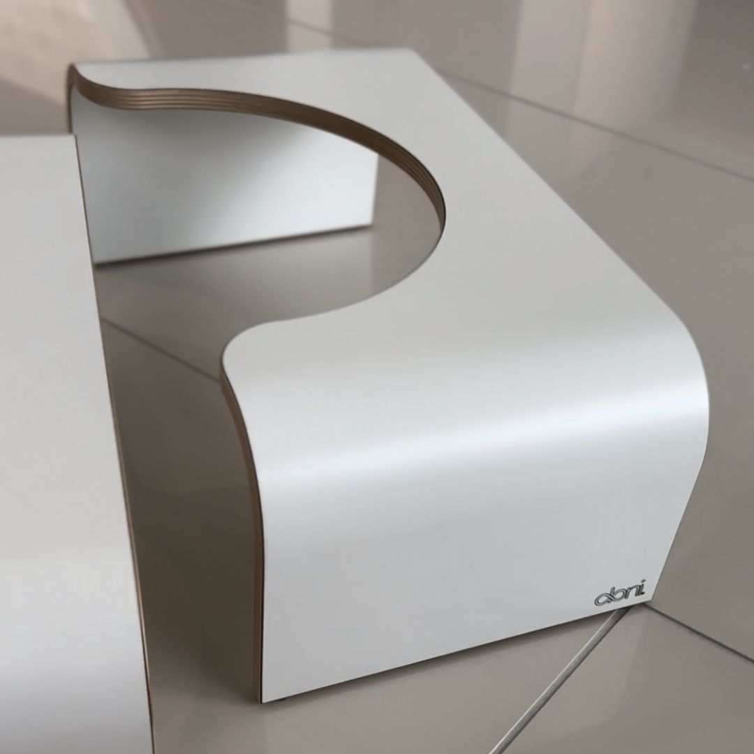A modern, white, C-shaped stool or bench with smooth curves sits on a glossy tiled floor. The minimalist object features layered wood edges and a sleek design. Object is made of wood with a white surface layer; visible edge grain along the open sides; no hardware or visible branding. Floor consists of large, polished tiles, color light grey or off-white. No people, hands, packaging, or labels in view. Shot is taken at an angle highlighting the full profile and floor reflection.