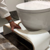 Oloni WC-jakkara (Kakkara); käsi säätää valkoista Squatty Potty -jakkaraa wc-istuimen edessä ruskeankuvioisella harmaalla matolla kylpyhuoneessa; jakkara muovia sileällä mattapinnalla ja kaarevin reunoin; kuvassa näkyy vain osa kädestä ja käsivarrestä; kylpyhuoneen lattiassa laatoitusta, ei pakkausta tai luettavaa etikettitekstiä näkyvissä.