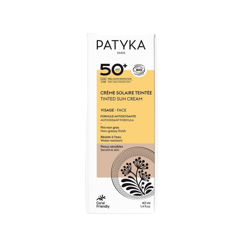 Patyka Tinted Face Sun Cream SPF50+; valkoinen laatikko Patyka-sävytettyä aurinkovoidetta kasvoille SPF 50+, mustin ja kultaisin yksityiskohdin, tekstitietoja ja kukkakuvitus etupuolella; pinta on mattapintainen; pakkaus on suorakulmainen terävin reunoin; typografia yhdistää lihavaa groteskia ja ohutta antiikvaa; etiketti mainitsee koralleja säästävän ja herkälle iholle sopivan; yksivärinen tausta, ei ihmisiä tai käsiä näkyvissä.