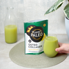 Planet Paleo Osteo Collagen; käsi pitelee läpinäkyvää juomalasia täynnä vihreää nestettä mattamustan suorakulmaisen pussin vieressä, merkitty ”Planet Paleo Osteo Collagen” lihavalla valkoisella tekstillä; pöydällä myös muovipullo läpinäkymättömällä valkoisella korkilla samalla merkinnällä sekä savinen ruukku, jossa lehtevä vihreä kasvi; kuva otettu sisätiloissa yläviistosta.