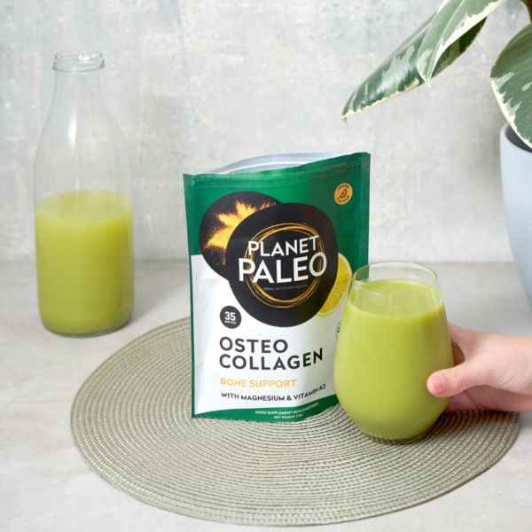 Planet Paleo Osteo Collagen; käsi pitelee läpinäkyvää juomalasia täynnä vihreää nestettä mattamustan suorakulmaisen pussin vieressä, merkitty ”Planet Paleo Osteo Collagen” lihavalla valkoisella tekstillä; pöydällä myös muovipullo läpinäkymättömällä valkoisella korkilla samalla merkinnällä sekä savinen ruukku, jossa lehtevä vihreä kasvi; kuva otettu sisätiloissa yläviistosta.