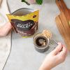 Planet Paleo Keto Coffee; henkilö pitelee mustaa joustavaa pakkausta tekstillä "Planet Paleo Keto Coffee" lihavoiduin valkoisin groteskikirjaimin ja kultaisin serif-kirjaimin; kollageenipitoista jauhetta kauhotaan läpinäkyvään lasimukiin, jossa kahvia; alusta vaaleasävyinen keittiötaso; vieressä suorakulmainen puinen leikkuulauta ja lehtevä ruukkukasvi; kuvassa näkyvät kädet, muki, pakkaus ja välineet sekä osa taustasta.