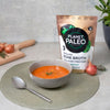 Planet Paleo Luomu Luuliemi Herbal Defence; kulho tomaattikeittoa yrtein vihreällä alustalla lusikan vieressä; taustalla mattamusta muovipakkaus tekstillä "Planet Paleo Organic Bone Broth collagen protein" lihavoiduin valkoisin ja kultaisin suuraakkosin; salottisipulia ja tuoreita yrttejä aseteltuna pienelle vaalealle puiselle leikkuulaudalle taustalla; ei ihmisiä tai käsiä; kuva otettu ylhäältäpäin.
