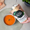 Planet Paleo Luomu Luuliemi Herbal Defence; käsi pitelee mattaharmaata ja mustaa uudelleensuljettavaa pussia tekstillä "Planet Paleo Organic Bone Broth Collagen Protein" punaisen tomaattikeittokulhon yläpuolella; alusliina kudottu, väriltään tan; vieressä puisella leikkuulaudalla lasiastia luonnonvalkoista kollageenijauhetta sekä pieni ruukkukasvi; tuotenimi selkeästi lihavoidulla valkoisella kirjasimella; käsi pitelee pussia keiton yläpuolella, viereen aseteltuna kasvi ja kollageenijauhe.