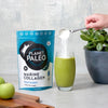 Planet Paleo Marine Collagen; käsi kaataa valkoista jauhetta valkoisesta kauhasta läpinäkyvään lasiin, jossa kirkkaanvihreää nestettä; vieressä tummanvihreä, mattapintainen Planet Paleo Marine Collagen -pussi pystyssä, etiketin teksti valkoisin groteskisuuraakkosin; kolme kiiltävää vihreää omenaa ja pieni ruukkukasvi mattaharmaassa ruukussa vaalealla, yhtenäisellä pöytätasolla; laaja kuvakulma ylhäältä.