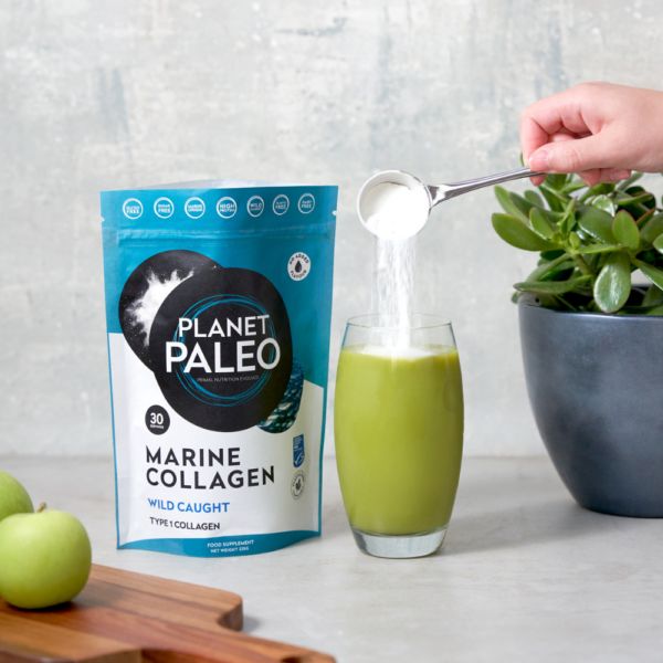 Planet Paleo Marine Collagen; käsi kaataa valkoista jauhetta valkoisesta kauhasta läpinäkyvään lasiin, jossa kirkkaanvihreää nestettä; vieressä tummanvihreä, mattapintainen Planet Paleo Marine Collagen -pussi pystyssä, etiketin teksti valkoisin groteskisuuraakkosin; kolme kiiltävää vihreää omenaa ja pieni ruukkukasvi mattaharmaassa ruukussa vaalealla, yhtenäisellä pöytätasolla; laaja kuvakulma ylhäältä.