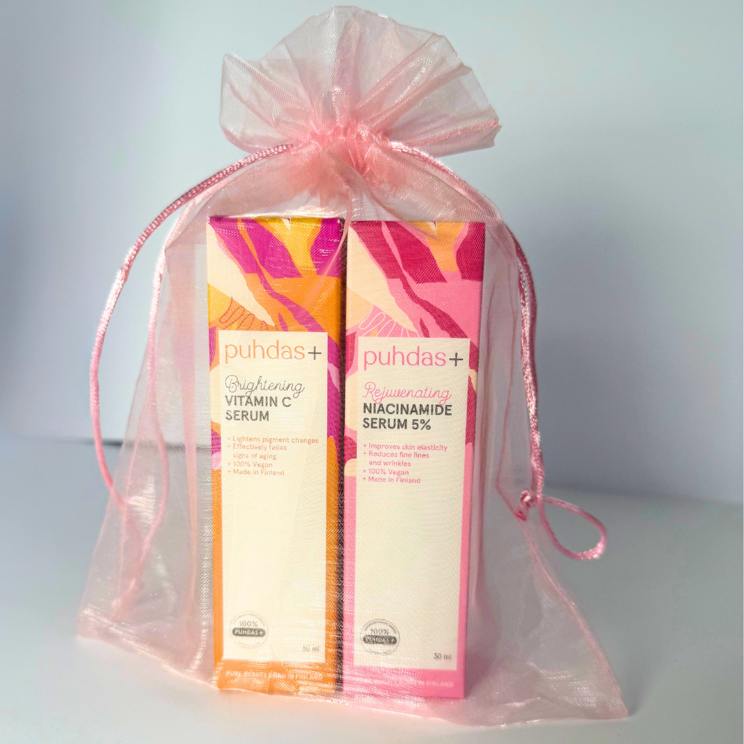 Puhdas Lahjapakkaus Niacinamide Serum & Vitamin C Serum; kaksi suorakaiteen muotoista ihonhoitoseerumipakkausta, merkitty Vitamin C ja Niacinamide, seisoo pystyssä läpinäkyvässä vaaleanpunaisessa organzapussissa satiininauhoin suljettuna; molemmat laatikot valkoisella taustalla painetuin tiedoin; fontit siistejä ja groteskeja; tausta tasainen vaaleanharmaa studiotyylisessä tuotekuvassa.