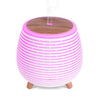 Puhdas+ Aroma Diffuser Ultrasonic 140 ml; pyöreä moderni eteerisen öljyn diffuusori vaaleanpunaisin ja valkoisin hehkuvin raidoin, puinen yläosa ja pienet puiset jalat; pinta mattainen valaistuin raidoin; ohut sumusuihku poistuu keskiaukosta; ei näkyviä etikettejä tai typografiaa; tuote neutraalilla tasaisella alustalla, ei ihmisiä tai käsiä; studiotyylinen tasaisesti valaistu kuva.