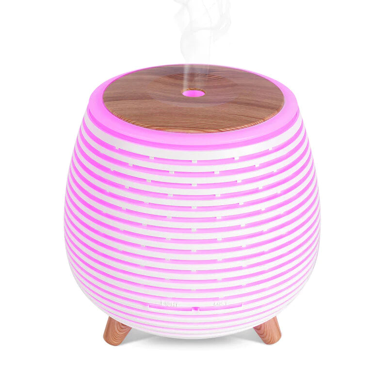 Puhdas Aroma Diffuser & Eteerinen Laventeliöljy Lahjapakkaus; pyöreä, moderni eteerisen öljyn diffuusori vaaleanpunaisin ja valkoisin raidallisin valoin, puinen yläosa ja kolme puujalkaa; aromiterapiahöyry näkyy tulevan yläosan aukosta; diffuusori mattapintaista muovia puunsyykuvioidulla kupolilla ja vastaavin puujaloin; ei näkyvää brändäystä tai etikettitekstiä; neutraali tausta, tuote esitetty eristettynä studiokuvana ilman ihmisiä tai käsiä.