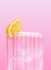 Korkea lasi täynnä vaaleanpunaista mocktailia ja jäätä, koristeltu sitruunalohkolla lasin reunalla, vaaleanpunaista liukuväritaustaa vasten. Kirkas lasi on lieriömäinen ja sileäreunainen, ja siinä näkyy jääpaloja nesteen läpi. Etikettiä tai typografiaa ei näy. Kuvassa on vain juoma; ihmisiä tai pakkausta ei näy.