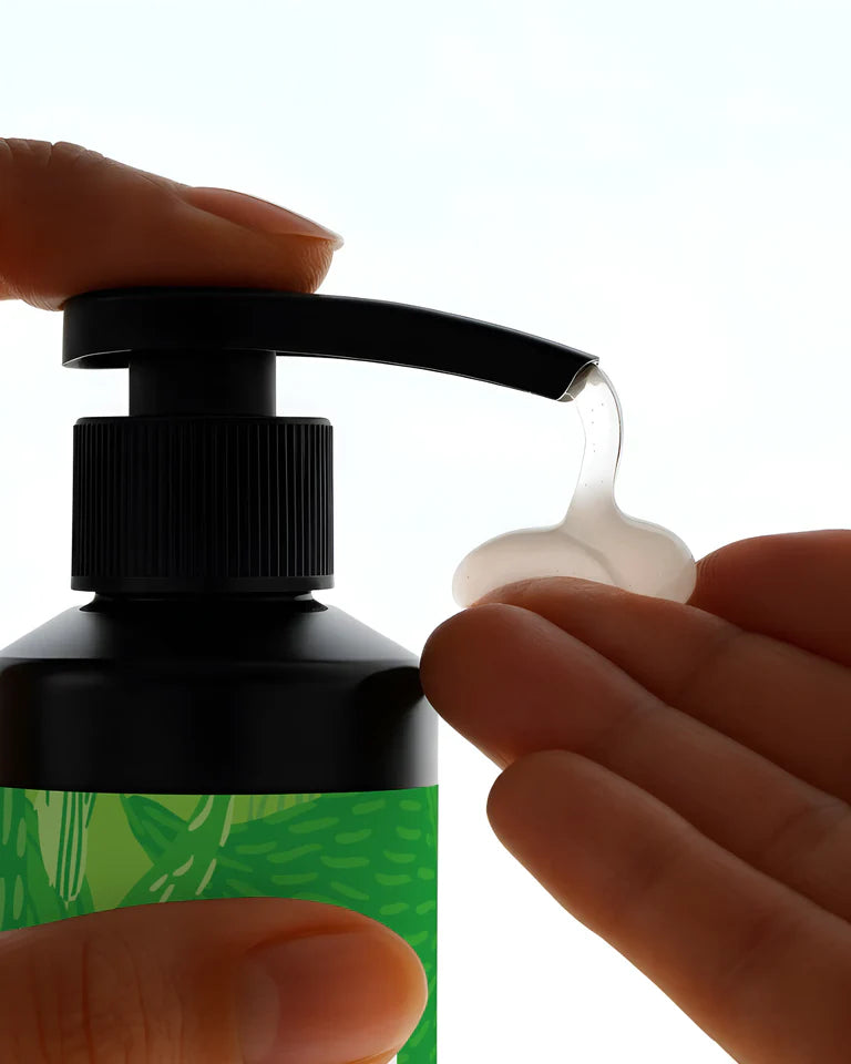 Puhdas+ Kofeiini Shampoo; käsi painaa pumppuannostelijaa vapauttaen kirkasta geeliä toiselle kädelle; tuote mustassa muovipullossa kiiltäväpinnalla ja kiinnitetyllä pumppukärjellä; suorakaiteen muotoinen vihreä etiketti kulmikkaalla kuviolla ja vähäisellä valkoisella groteskitekstillä; tausta tasaisesti valaistu vaalea.