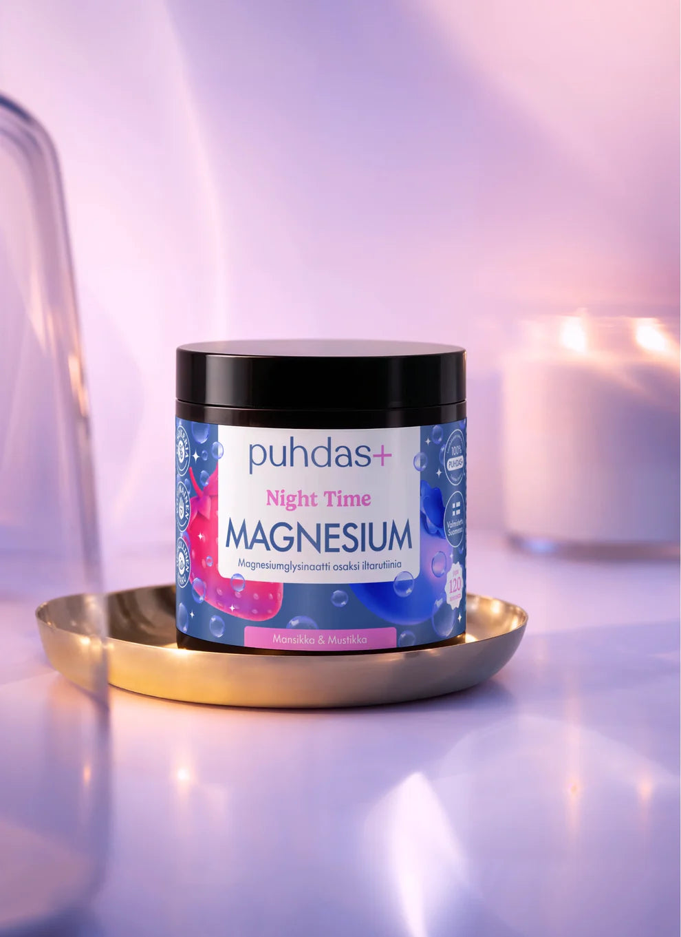 Lieriömäinen purkki, jossa lukee Puhdas+ Night Time Magnesium powder, on aseteltu pyöreälle, heijastavalle kultaiselle lautaselle. Astia on valkoinen ja mattapintainen, ja siinä on sininen ja vaaleanpunainen etiketti pienin marjakuvituksin. Pääteviivaton typografia näyttää tuotenimen ja kuvauksen. Taustalla on lämminsävyinen sumea pinta, jolla näkyy palava kynttilä. Ihmisiä tai käsiä ei näy kuvassa.