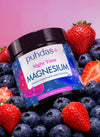 Valkoinen muovinen lieriömäinen Puhdas+ Night Time Magnesium -purkki on aseteltu tuoreiden mansikoiden ja mustikoiden joukkoon tasaiselle pinnalle. Etiketti on sininen ja violetti vaaleanpunaisin viivoin, selkeällä pääteviivattomalla typografialla ja suomenkielisellä tekstillä. Valaistus on tasainen, tausta epätarkka. Ihmisiä tai käsiä ei näy.