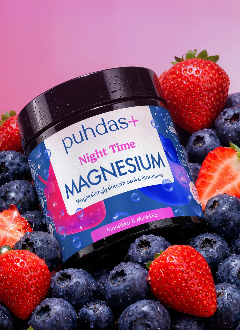 Valkoinen muovinen lieriömäinen Puhdas+ Night Time Magnesium -purkki on aseteltu tuoreiden mansikoiden ja mustikoiden joukkoon tasaiselle pinnalle. Etiketti on sininen ja violetti vaaleanpunaisin viivoin, selkeällä pääteviivattomalla typografialla ja suomenkielisellä tekstillä. Valaistus on tasainen, tausta epätarkka. Ihmisiä tai käsiä ei näy.