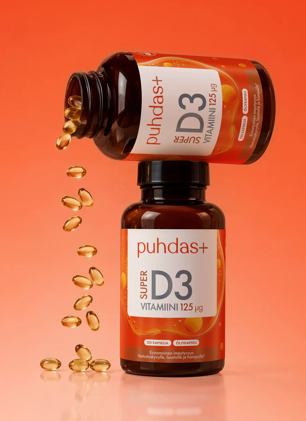 Kaksi Puhdas+ Super D3 Vitamiini -pulloa, toinen pystyssä ja toinen kaatuneena, pehmytkapseleiden valuessa ulos oranssia hyvinvointihenkistä taustaa vasten. Molemmat pullot ovat valkoista muovia, lieriömäisiä ja mattapintaisia. Etiketeissä lukee Puhdas+ Super D3 Vitamiini pääteviivattomalla fontilla. Ihmisiä tai käsiä ei näy; kuva on suora tuotekuva.