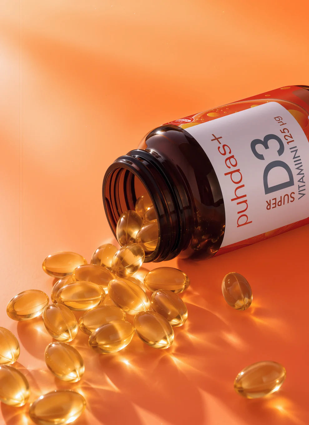 Pullo, jossa lukee SUPER D3 VITAMIN D3 125µg, makaa kyljellään ja siitä valuu soikeita, läpikuultavia kultaisia kapseleita oranssille mattapinnalle. Muovipullo on valkoinen rungoltaan, ja siinä on lihavoitua pääteviivatonta mustaa ja oranssia etikettitekstiä. Ihmisiä tai käsiä ei näy. Kuva on lähikuva, jossa näkyy vain tuote ja kapselit yksiväristä taustaa vasten.