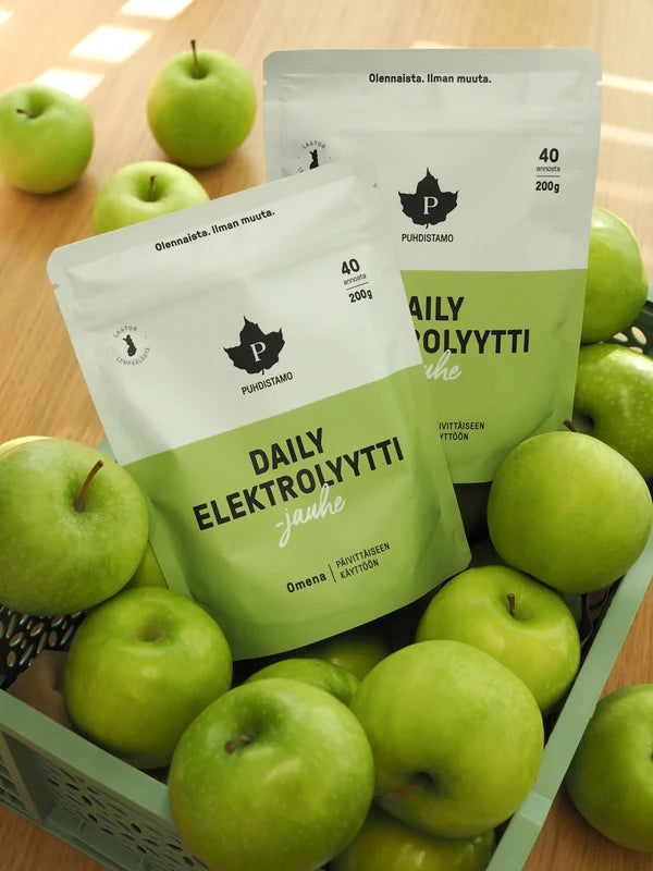 Kaksi valkoista mattapintaista pussia tekstillä 'Daily Elektrolyytti' lihavoidulla pääteviivattomalla fontilla seisovat pystyssä vaaleassa punotussa korissa täynnä vihreitä omenoita. Taustalla näkyy lisää vihreitä omenoita puisella alustalla. Ihmisiä tai käsiä ei näy, eikä muita esineitä tai tuotteita ole näkyvissä.