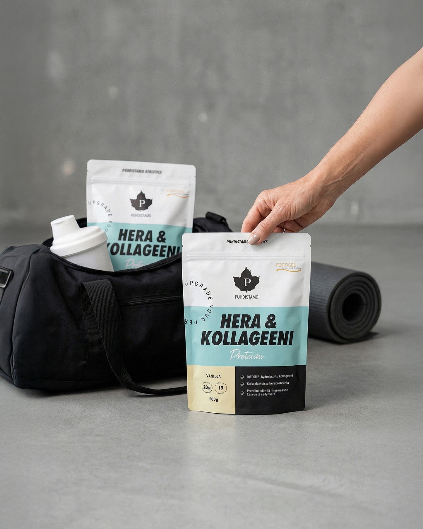 Puhdistamo Hera & Kollageeni Vanilja; käsi nostaa matan pussin tekstillä ”Hera & Kollageeni Proteiin” mustan vetoketjullisen kuntosalilaukun, valkoisen sylinterin muotoisen shakerin ja sinisen rullatun joogamaton läheisyydessä sileällä harmaalla lattialla; etiketin teksti selkeää groteskia; muita ihmisiä ei näy; esineet ryhmiteltynä lähelle toisiaan, kuva yläviistosta.