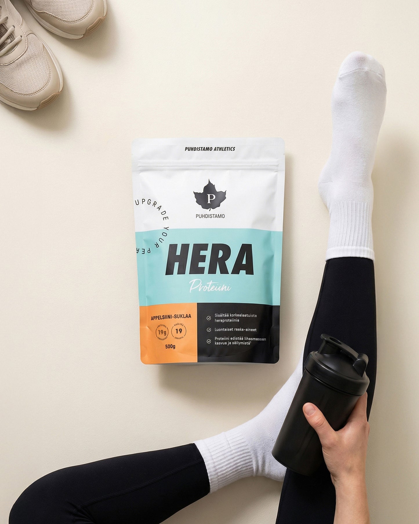 Puhdistamo Heraproteiini Appelsiini-Suklaa; henkilö valkoisissa sukissa ja mustissa legginseissä istuu jalat ristässä, kädessä musta matta muovinen shakerpullo HERA-proteiinipakkauksen vierellä beigellä tasaisella taustalla; proteiinipakkauksen groteskikirjasin lihavoitu ja moderni, tekstinä ”HERA”; kuvan kulmassa osittain näkyvissä beige kangas- tai mokkatennari.