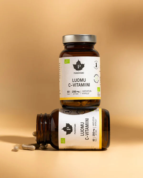 Puhdistamo Luomu C-vitamiini; kaksi ruskeaa lisäravinnepulloa, merkitty Luomu C-Vitamiini, on pinottu beigelle pinnalle, toinen pystyssä ja toinen makuulla; vieressä kaksi kapselia ja tabletti; molemmissa pulloissa mattapintainen muovi ja sylinterinmuoto; etiketissä selkeää mustaa ja vihreää groteskitekstiä; tausta vaaleanbeige, ei ihmisiä tai muita esineitä näkyvissä.