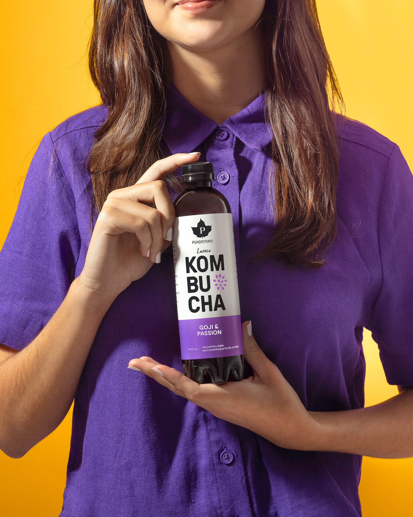 Violettipaitainen nainen pitelee läpinäkyvää, lieriömäistä lasipulloa mattaetiketillä, jossa lukee Kombucha ja maut Goji ja Passionfruit groteskikirjasimella; pullon sisällä oleva neste on meripihkanvärinen; tausta kirkkaan keltainen; keskikokoinen lähikuva keskittyy pulloon ja naisen yläkropan yläosaan.