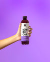 Käsi pitelee lieriömäistä, läpinäkyvää pulloa, jossa violettia kombuchaa; etiketissä lukee KOMBUCHA Cool & Passion suurin, valkoisin groteskikirjaimin; pullossa hopeanvärinen korkki ja sileä kiiltäväpinta; tausta yhtenäisen violetti ilman muita esineitä tai tekstuureja; kuva otettu edestä ja rajattu tiiviisti.