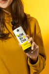 Puhdistamo Luomu Kombucha Inkivääri-Sitruuna; keltaiseen collegepaitaan pukeutunut nainen pitelee kirkasta lasipulloa, jossa inkivääri-sitruunakombuchaa; pullo sylinterimäinen, etiketissä keltaista ja valkoista väriä sekä lihavoitua, modernia typografiaa ja tuotenimi luettavissa; etiketin pinta mattaa; tausta yhtenäisen kirkkaan keltainen, tasaisesti valaistu.