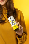 Puhdistamo Luomu Kombucha Inkivääri-Sitruuna; keltaiseen collegepaitaan pukeutunut nainen pitelee kirkasta lasipulloa, jossa inkivääri-sitruunakombuchaa; pullo sylinterimäinen, etiketissä keltaista ja valkoista väriä sekä lihavoitua, modernia typografiaa ja tuotenimi luettavissa; etiketin pinta mattaa; tausta yhtenäisen kirkkaan keltainen, tasaisesti valaistu.