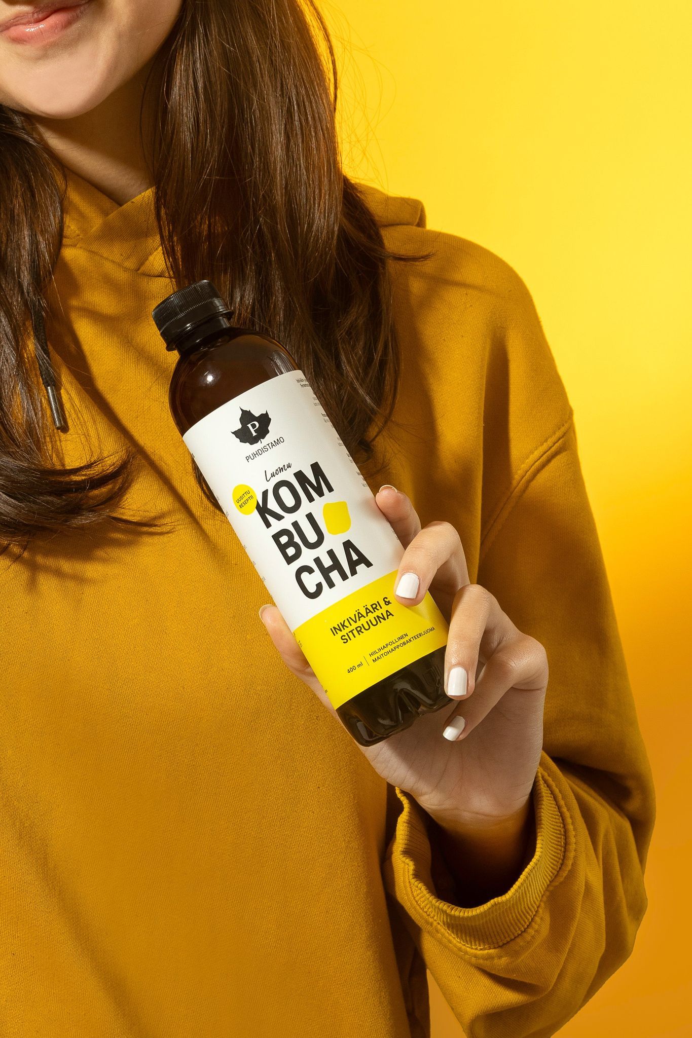 Puhdistamo Luomu Kombucha Inkivääri-Sitruuna; keltaiseen collegepaitaan pukeutunut nainen pitelee kirkasta lasipulloa, jossa inkivääri-sitruunakombuchaa; pullo sylinterimäinen, etiketissä keltaista ja valkoista väriä sekä lihavoitua, modernia typografiaa ja tuotenimi luettavissa; etiketin pinta mattaa; tausta yhtenäisen kirkkaan keltainen, tasaisesti valaistu.