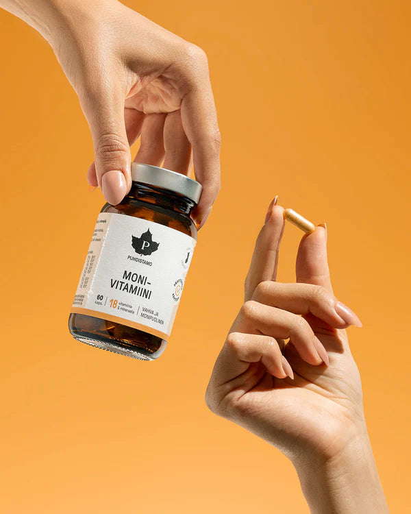 Puhdistamo Monivitamiini; käsi pitelee muovipulloa, jossa selkeä "Multivitamin"-etiketti groteskikirjasimella; toinen käsi esittelee yhden pitkulaisen kapselin; pullo on sylinterinmuotoinen ja mattapintainen; tausta yhtenäisen oranssi; kuvassa näkyy vain kädet, ei muita esineitä tai ympäristöä; lähikuva.