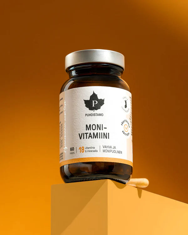 Puhdistamo Monivitamiini; ruskea lasipurkki seisoo keltaisella jalustalla oranssia taustaa vasten, vieressä vitamiinipitoisia kapseleita; purkki on sylinterinmuotoinen mustalla kierrekorkilla ja mattapinnalla; etiketissä groteskikirjasin esittää tuotenimen ja listaa tärkeimmät vitamiinit ja kivennäisaineet sekä vaahterapuun lehtilogon; ei ihmisiä tai käsiä näkyvissä.