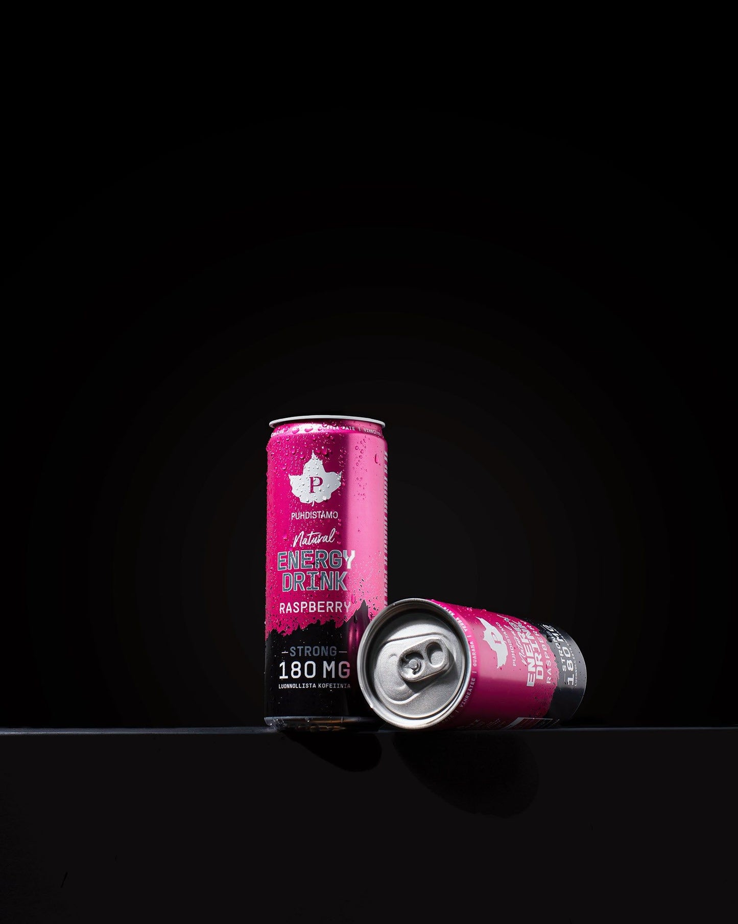 Two pink cans of energy drink rest on a black surface, one upright and one lying down. They are standard aluminum cans with a glossy finish and covered in condensation. The design features bold, modern typography displaying “raspberry flavor” and “180 mg caffeine” on the label. No people or hands present. The shot is against a solid black background.