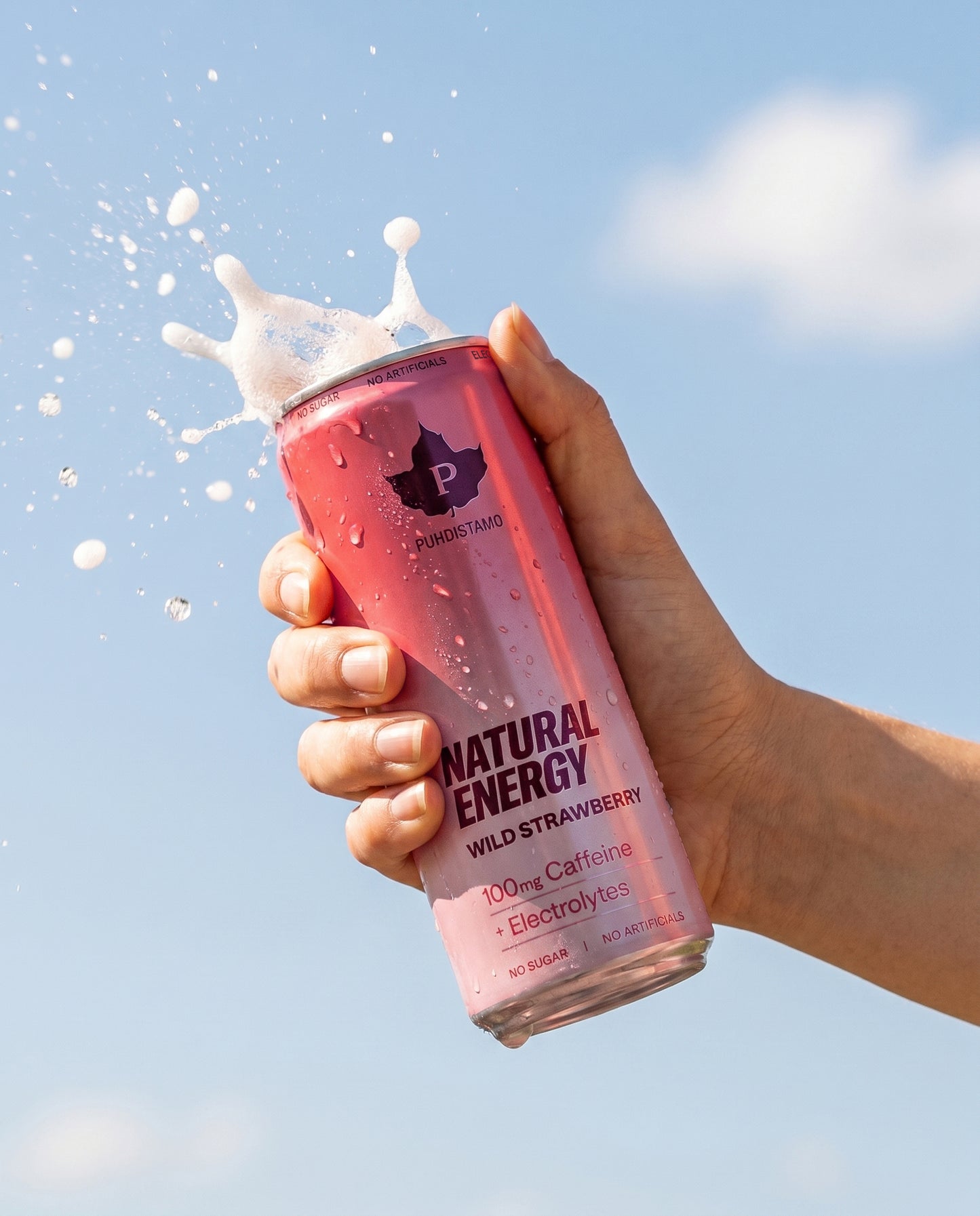 Puhdistamo Natural Energy Wild Strawberry: käsi pitelee mattapintaista pinkkiä alumiinitölkkiä tekstillä "Natural Energy Wild Strawberry" lihavoidulla, groteskilla valkoisella ja punaisella kirjasimella; valkoista nestettä suihkuaa avatusta tölkin yläosasta; tausta on kirkas, selkeä sininen taivas; kuva on lähikuva keskittyen tölkkiin ja käteen; kuvassa näkyy vain yksi ihmiskäsi, joka pitelee tölkkiä alaosastaan.