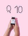 Two hands hold a bottle of supplements against a pink background. The bottle is cylindrical with a white label and white cap, matte surface finish. Dark capsules are neatly arranged above the bottle to form the letters “Q 10”. No other visible text is legible on the label. The shot is tightly framed, focusing only on hands, bottle, and capsules.