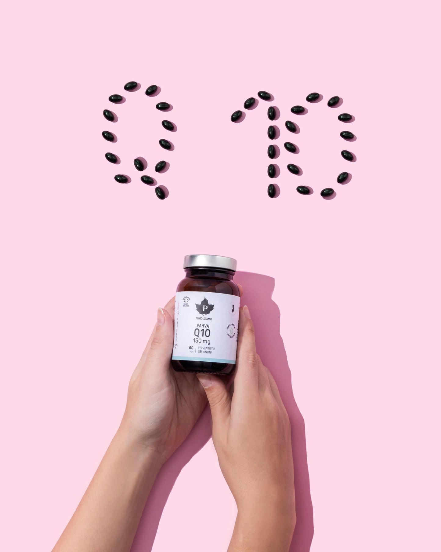 Two hands hold a bottle of supplements against a pink background. The bottle is cylindrical with a white label and white cap, matte surface finish. Dark capsules are neatly arranged above the bottle to form the letters “Q 10”. No other visible text is legible on the label. The shot is tightly framed, focusing only on hands, bottle, and capsules.