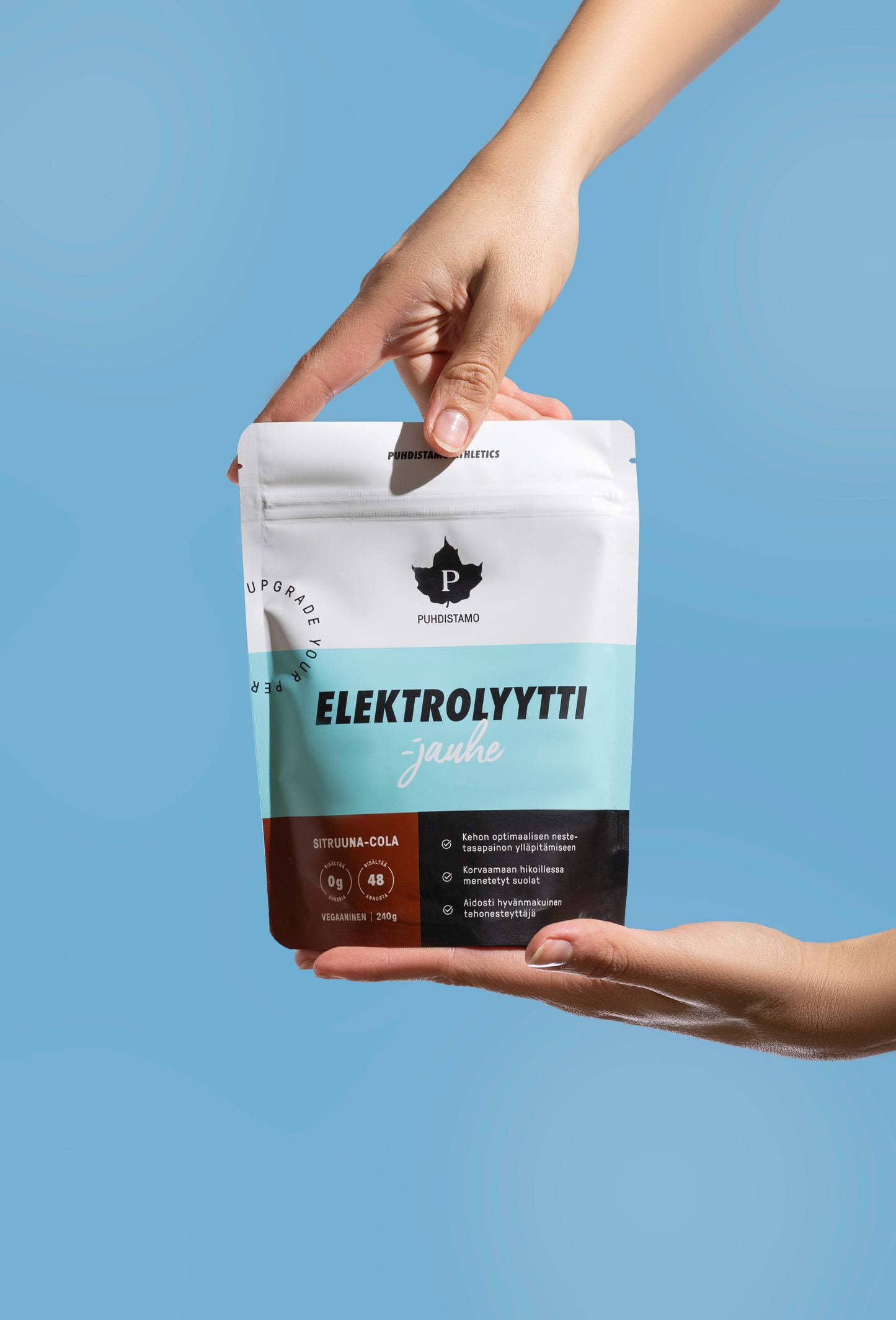 Puhdistamo Elektrolyyttijauhe Sitruuna-Cola; kaksi kättä pitelee valkoista ja sinistä uudelleensuljettavaa pussia tekstillä ”Elektrolyytti jauhe” tasaista vaaleansinistä taustaa vasten; mattapintainen pussi suorakaiteen muotoinen pyöristetyin kulmin, selkeää mustaa ja sinistä tekstiä, avaintuotetietolohkoja ja lihavoitu logo yläosassa; kaikki näkyvä sisältö liittyy elektrolyyttijauheeseen.