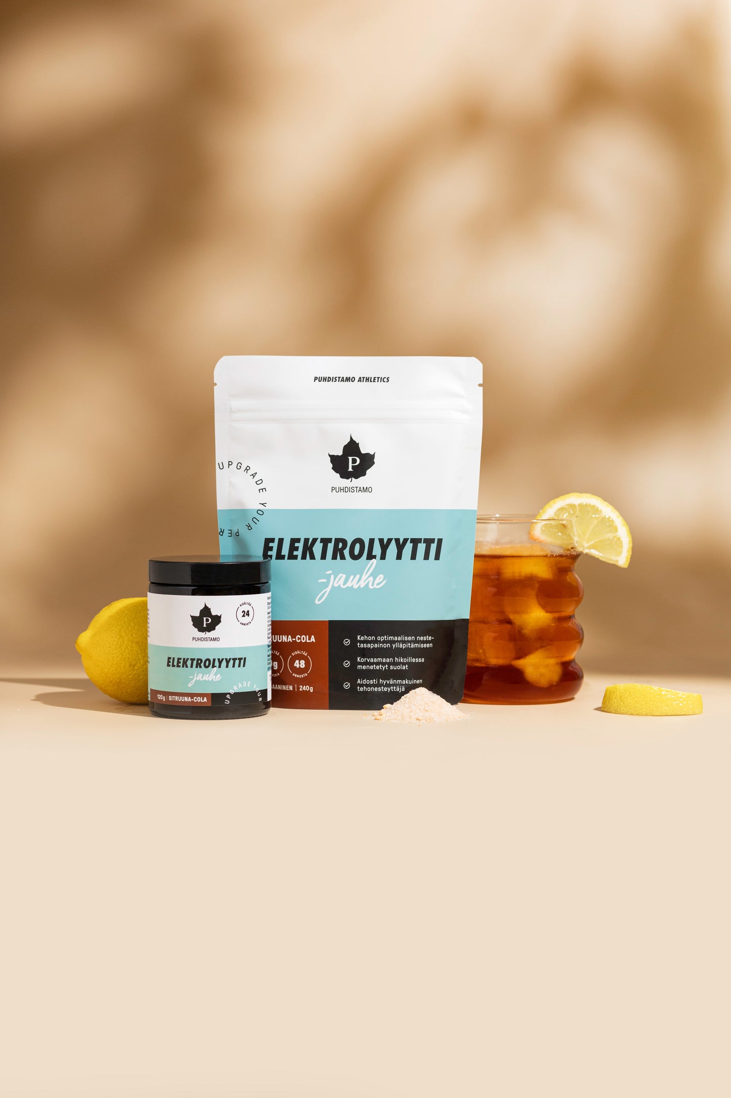 Puhdistamo Elektrolyyttijauhe Sitruuna-Cola; kaksi Elektrolyytti-jauheen astiaa, purkki ja pussi, esillä yhdessä jäätee-purkin, sitruunaviipaleen, irtonaisen elektrolyyttijauheen ja kahden sitruunalohkon kera beigellä pinnalla pehmeästi sumeaa taustaa vasten; purkki lieriömäinen mattavalkoisella pinnalla, merkitty ”Elektrolyytti” suurella tikkukirjasimella; pussi samansävyinen ja -typografinen; jäätee kirkkaassa lasipurkissa, jossa näkyvissä kondenssia; ei käsiä tai ihmisiä näkyvissä.