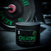 Puls Creatine, maku Pear; musta mattapintainen kreatiinimonohydraattijauhepurkki aseteltuna kuntosalivälineiden yhteyteen; lieriömäinen pakkaus kierrekannella; valkoinen etiketti lihavoiduin modernein groteskikirjaimin: zero sugar, 200g, 30 annosta puhdasta kreatiinimonohydraattia; taustalla epätarkka painolevy; ei ihmisiä tai käsiä.