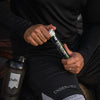 Puls Electrolyte Pro Poretabletti Päärynä: mustissa urheiluvaatteissa oleva henkilö pitelee putkiloa tekstillä "Electrolyte Pro" selkeällä modernilla groteskikirjasimella; musta vesipullo mattapinnalla ja valkoisella etiketillä on lähellä maassa; henkilö istuu ulkona betonilla, osittain suorassa auringonvalossa; kuvassa näkyy vain alavartalo ja käsi.