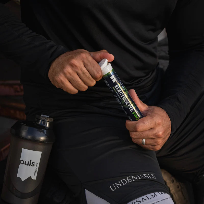 Puls Electrolyte Pro Poretabletti Päärynä: mustissa urheiluvaatteissa oleva henkilö pitelee putkiloa tekstillä "Electrolyte Pro" selkeällä modernilla groteskikirjasimella; musta vesipullo mattapinnalla ja valkoisella etiketillä on lähellä maassa; henkilö istuu ulkona betonilla, osittain suorassa auringonvalossa; kuvassa näkyy vain alavartalo ja käsi.