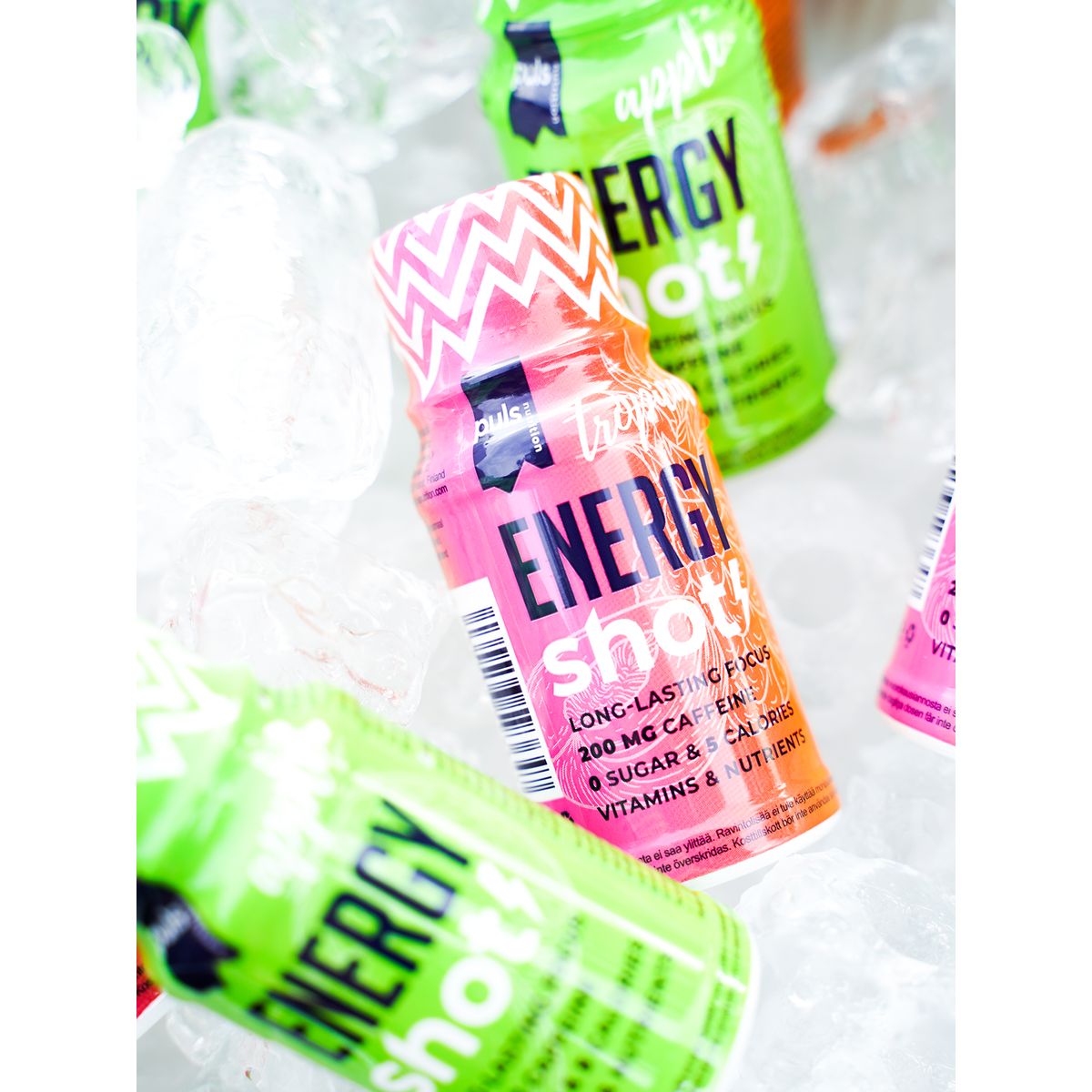 Puls Energy Shot Omena; värikkäitä energiajuomapulloja vihreässä ja pinkissä sävyssä aseteltuna kirkkaiden jääpalojen keskelle tasaiselle alustalle; pullot pieniä sylinterinmuotoisia muovipulloja kiiltäväpintaisin etiketein; pinkki pullo keskellä tekstillä "Energy Shot", kofeiinipitoisuus ja tärkeät vitamiinit lihavoidulla groteskikirjasimella; ei ihmisiä tai käsiä, lähikuva.