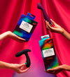 Three hands hold colorful vibrator boxes and sex toys against a vibrant red and pink fabric backdrop. Products include silicone vibrators in bright colors, with smooth or slightly curved shapes; both glossy and matte finishes are present. Packaging features bold sans-serif typography; legible text includes “MORE PLEASURE” and “ON SPOT STIMULATOR.” Shot is close-up, focusing on products.