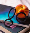 A bitten peach rests on a stack of books with three sleek black silicone rings arranged in front. The books have visible spines but no legible text. The rings are circular, solid black, and matte finished, positioned side by side in the foreground. No packaging is present. The background is neutral and out of focus; no people or hands are shown.