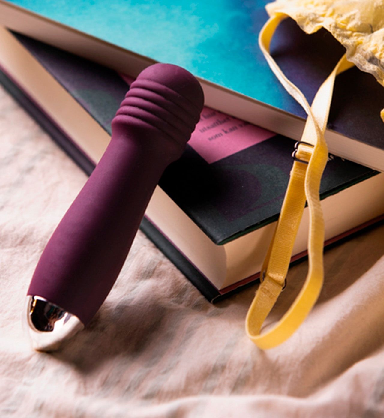 A purple personal massager with a smooth, matte finish is placed next to two stacked hardcover books; the covers and spines of the books are not fully visible and any text is obscured. A yellow lace bra with thin straps is draped nearby. All items rest on a soft, light-colored fabric surface in a flat lay arrangement.